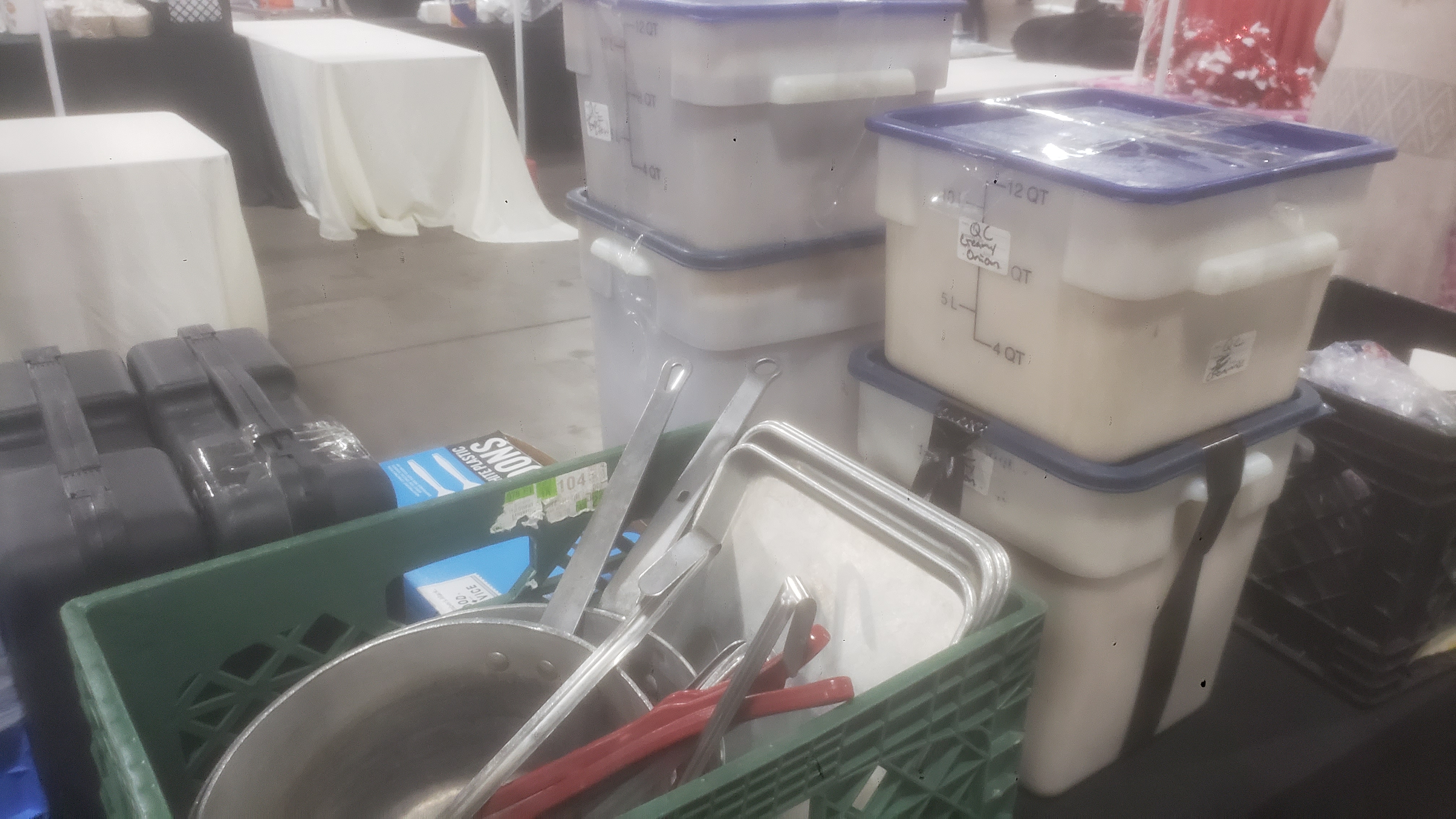Commercial food storage containers of soup and various supplies such as pans on a table with tables in the background.